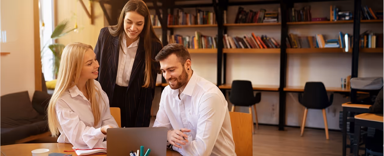 Three colleagues smiling and collaborating around a laptop in a modern office with bookshelves.