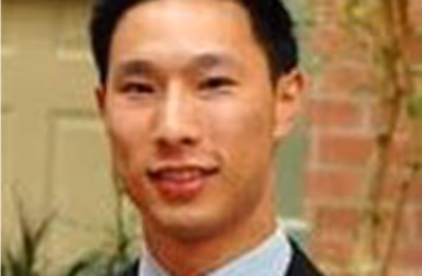 Smiling man in a suit and tie standing outdoors near plants and a brick wall.