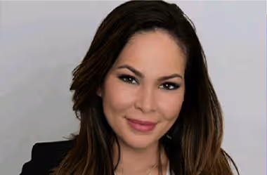 Woman with long dark hair and light makeup smiling against a plain light background.