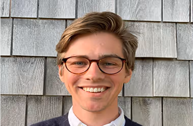 Smiling young man with glasses and brown hair standing in front of gray wooden shingles.