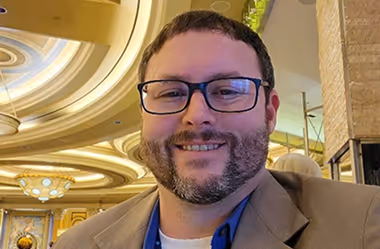 Smiling man with glasses and beard wearing a brown jacket in an elegant indoor setting.