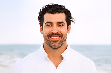 Smiling man with dark hair and beard wearing a white shirt near the ocean on a clear day.