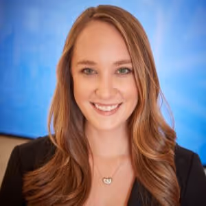 Smiling woman with long brown hair and green eyes in a black blazer against a blue background.