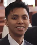 Young man with short black hair wearing a suit jacket and white shirt, smiling indoors.