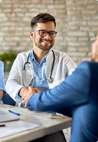 Smiling male doctor shaking hands with a patient across a desk in a consultation room.