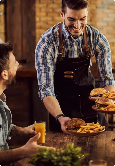 Smiling waiter serving plates with burgers and fries to a seated customer holding a glass of orange juice.