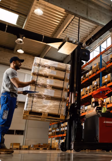 Worker in warehouse lifting wrapped pallets with forklift near tall shelves of boxes.