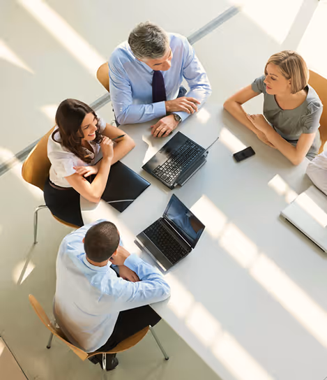 Four businesspeople sitting around table with laptops and documents engaged in discussion.