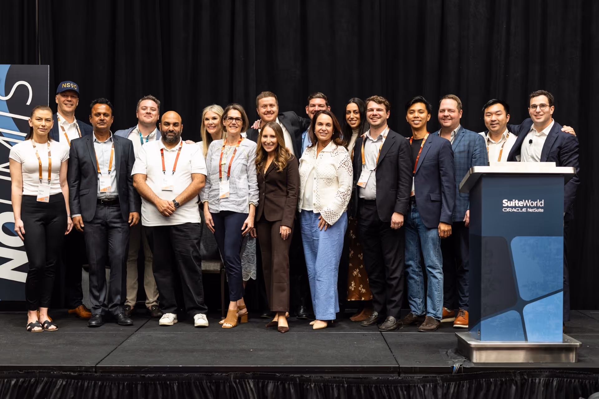 Group of sixteen people standing on stage at SuiteWorld Oracle NetSuite event with a podium on the right.