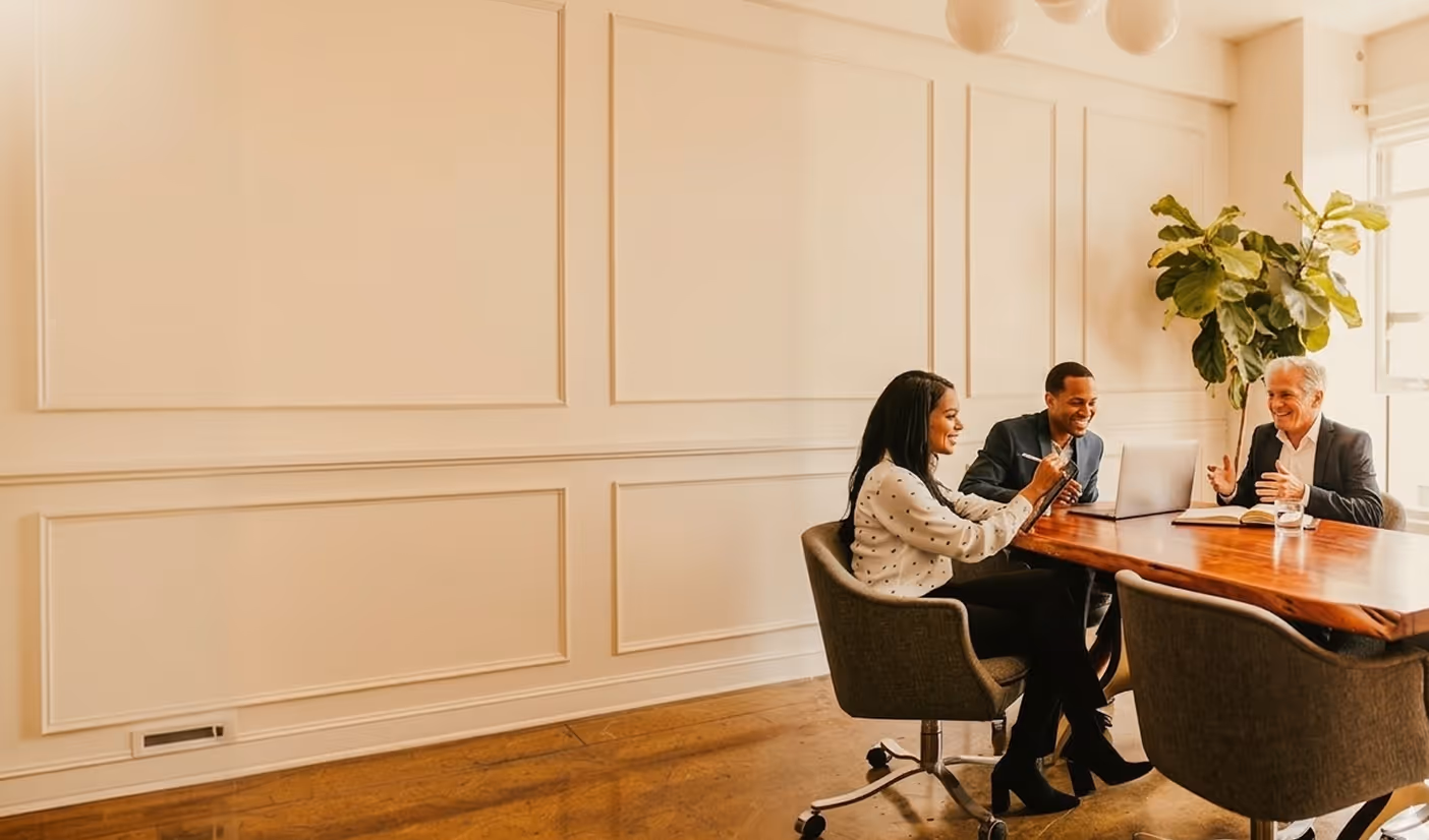 Three professionals sitting around a wooden table in a meeting room, discussing work with laptops and notes.