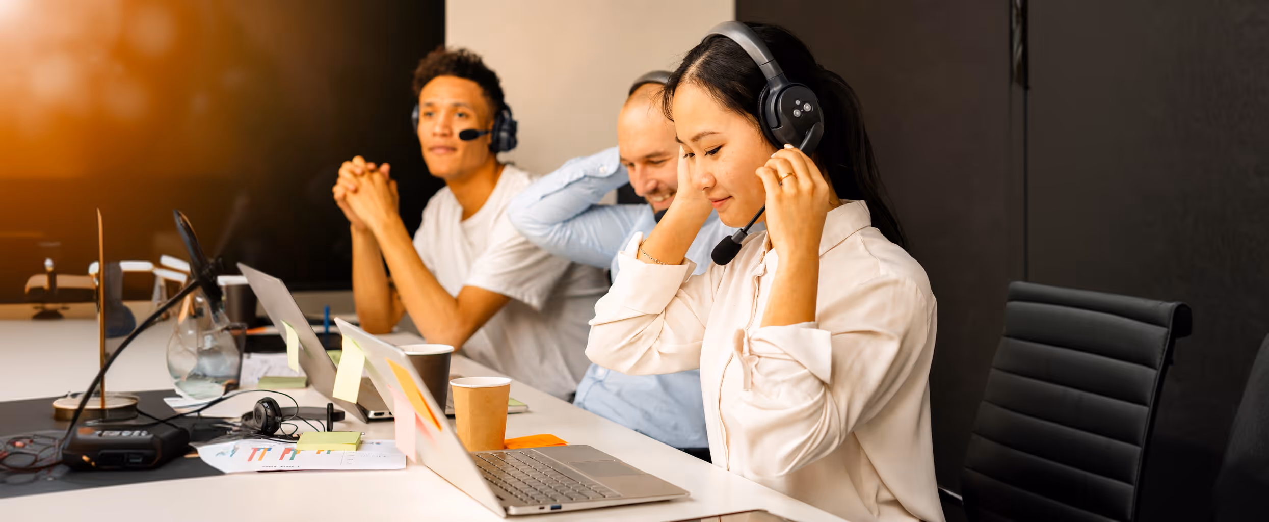 Three call center agents wearing headsets working at desks with laptops and coffee cups.