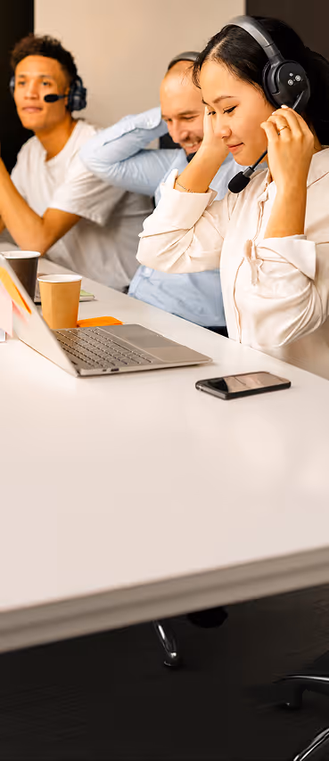 Three diverse call center agents with headsets working at a desk with laptop and smartphone.