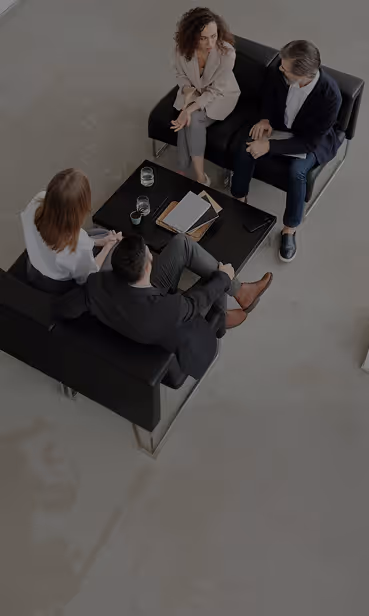 Four businesspeople in formal wear sitting on black sofas around a coffee table with documents and glasses.