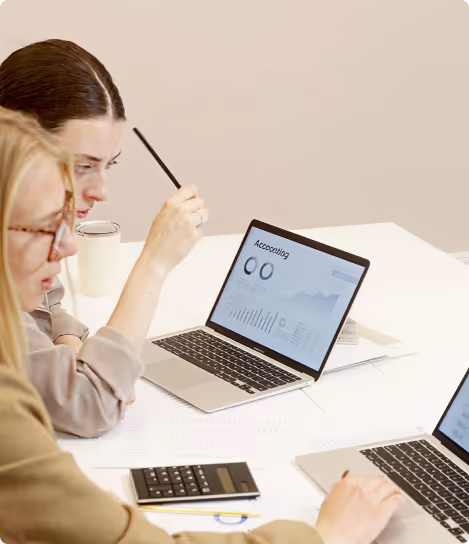 Two women working on laptops with accounting charts and a calculator on the desk.