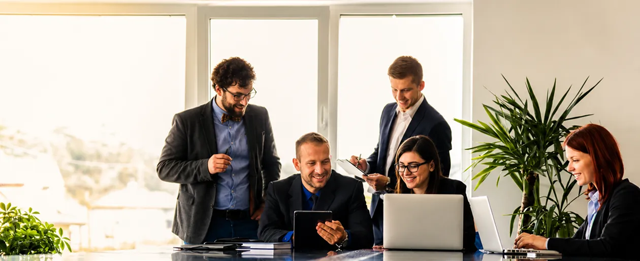Five coworkers smiling and working together at a table with laptops and documents in an office.