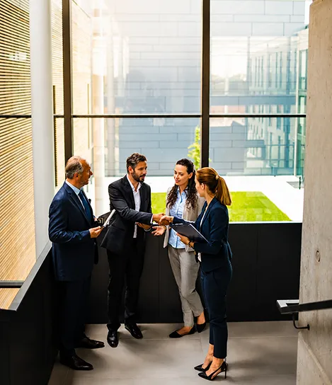 Four business professionals in a modern office hallway shaking hands and holding documents.