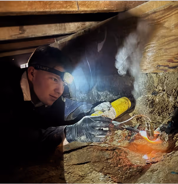 Person uses a blowtorch to heat a pipe connection in a dim crawlspace.