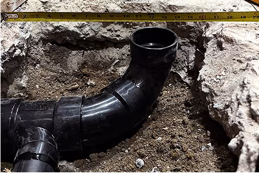 Measuring tape stretched above a recessed black plumbing pipe joint in dirt.