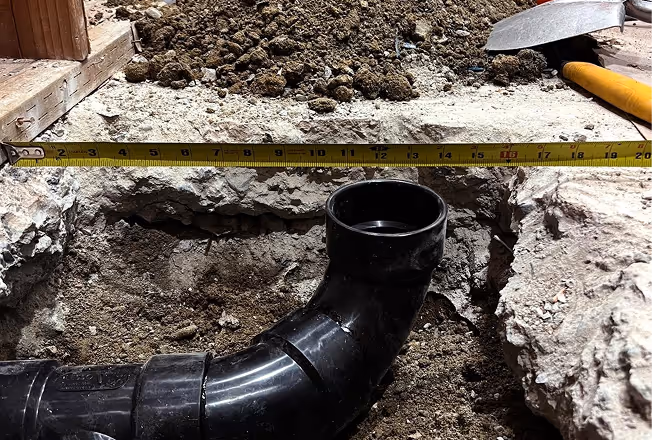 Measuring tape stretched above a recessed black plumbing pipe joint in dirt.