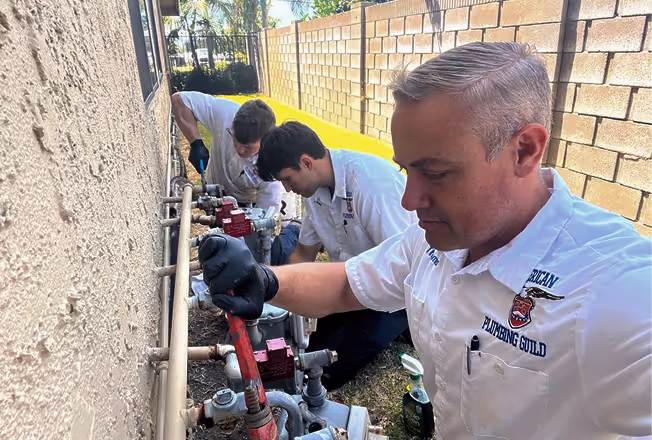 Three plumbers working on outdoor water meters along a house wall.