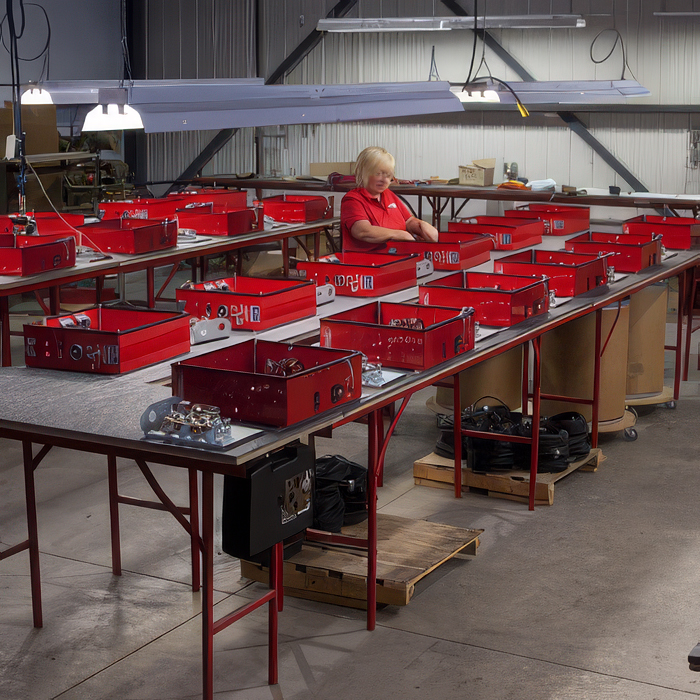Une femme travaille dans un atelier avec plusieurs caisses rouges sur des tables longues et hautes.