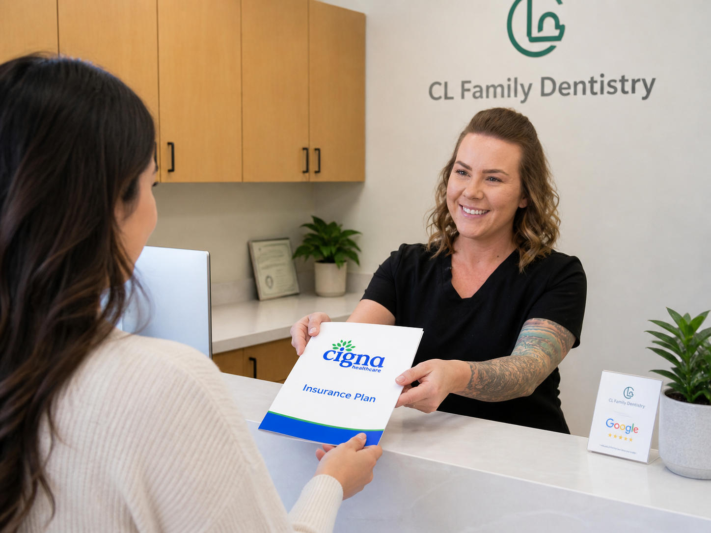 A friendly dental office assistant in black scrubs stands behind a clean, modern reception desk, smiling as she hands an insurance plan document to a patient. The well lit space features light wood cabinetry, indoor plants, and a welcoming, professional at