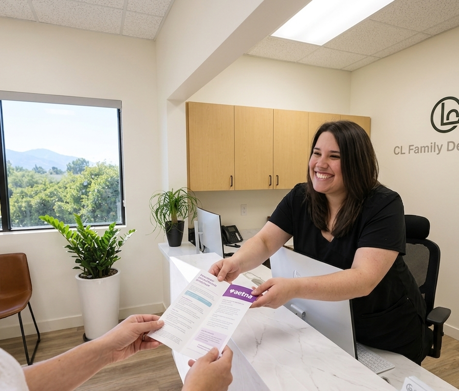 A friendly dental office assistant with a genuine smile presents an open trifold Aetna insurance brochure to a patient’s hands across a marble reception desk. The modern office background features a 'CL Family Dentistry' logo on the wall, a large window wi