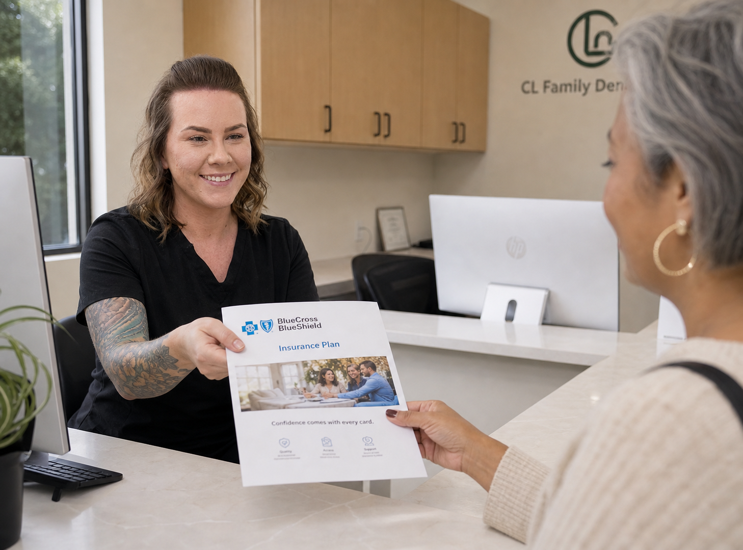 A friendly office assistant in a modern dental reception area hands a Blue Cross Blue Shield insurance plan document to a seated patient across the desk, both smiling in a bright, welcoming environment.
