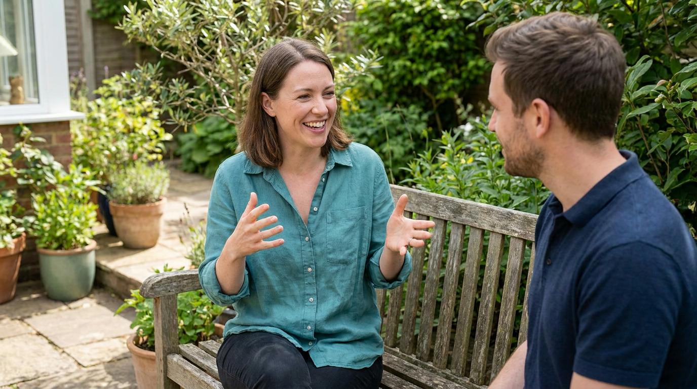 Zoe, wearing a teal shirt, sits on a wooden bench in a garden, smiling and gesturing while talking to Joshua, who is listening intently.