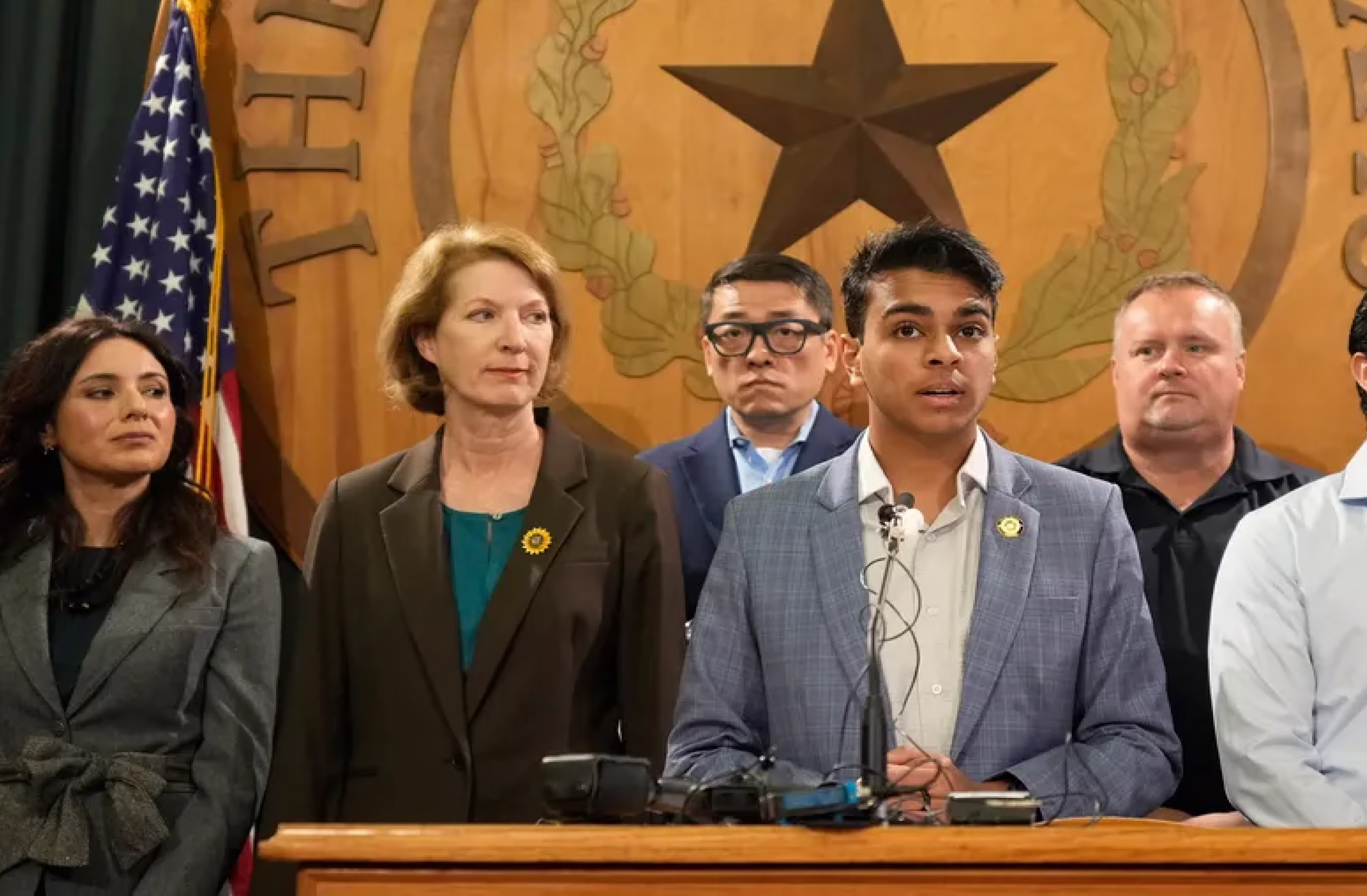 Five people standing at a podium with microphones, an American flag, and a large wooden emblem with a star in the background.