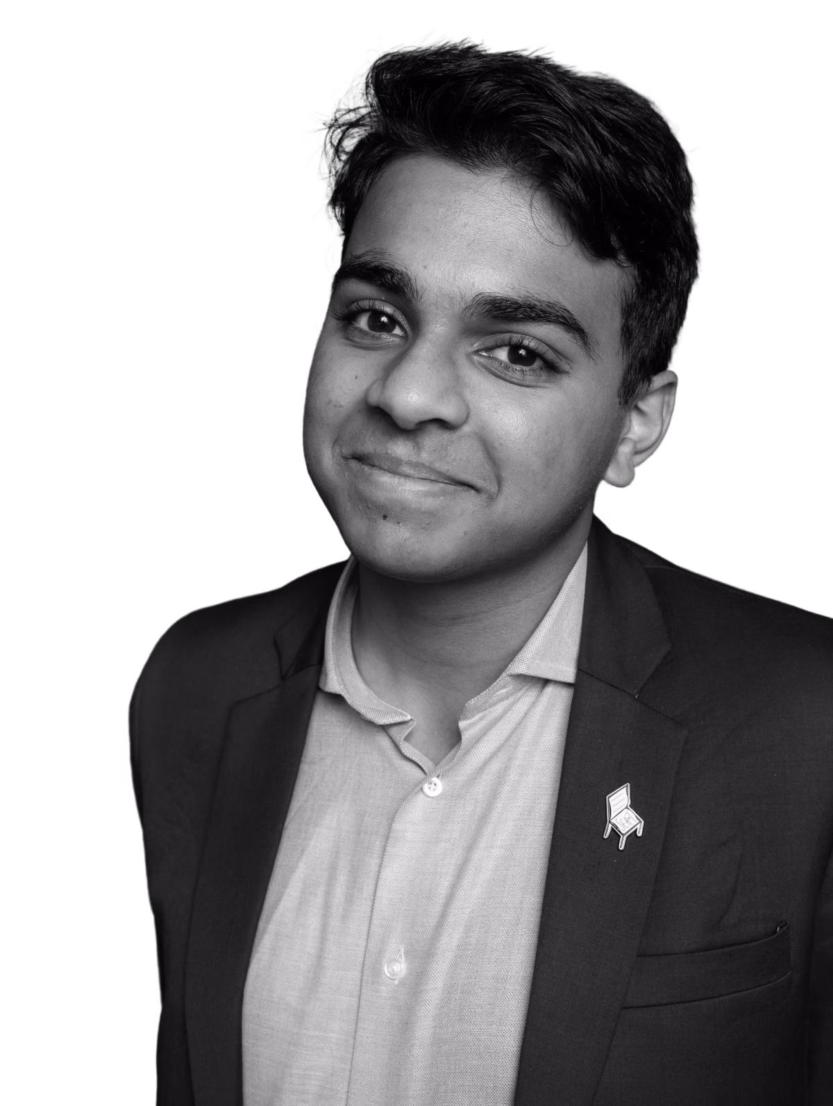 Smiling young man wearing a blazer with a pin of a chair on the lapel and a buttoned-up shirt.