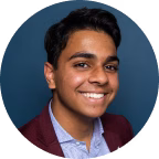 Smiling young man with dark hair wearing a burgundy blazer and light blue shirt against a dark blue background.