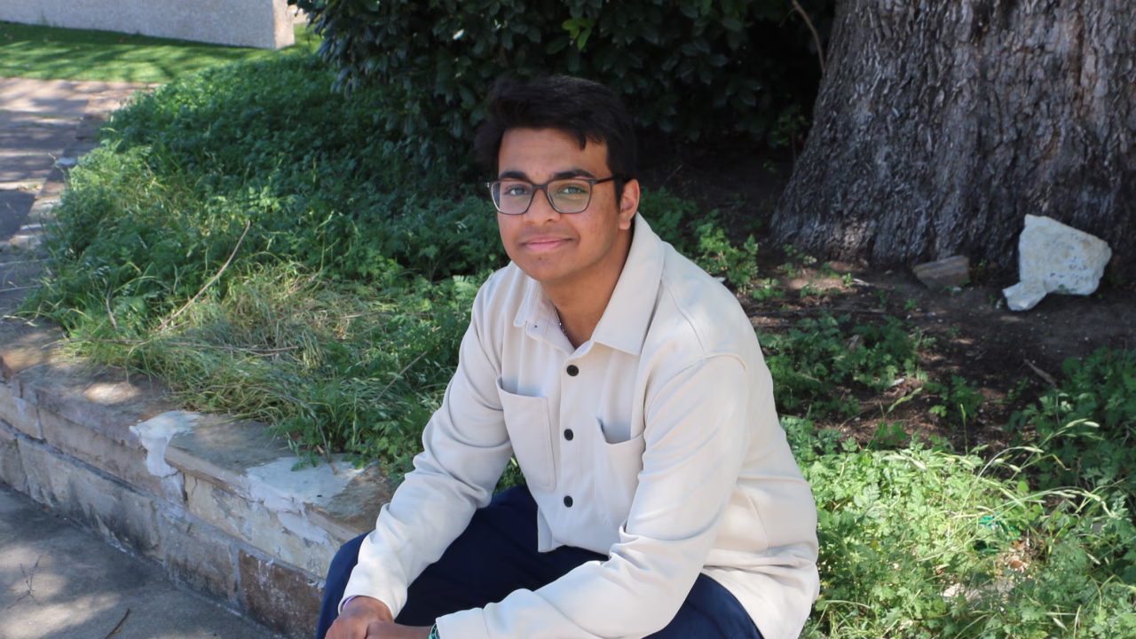 Young man wearing glasses and a light-colored shirt sitting outdoors near a tree and greenery.
