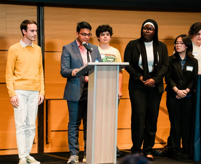 Group of diverse young adults standing on stage with one person speaking at a podium microphone.