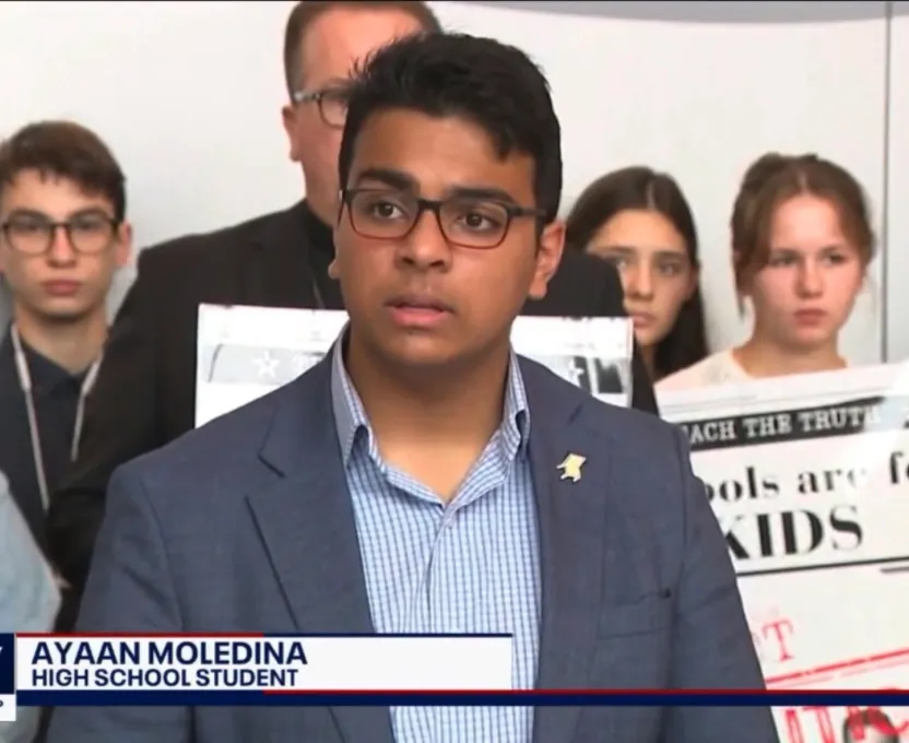 High school student Ayaan Moledina speaking at a podium with people holding protest signs behind him.