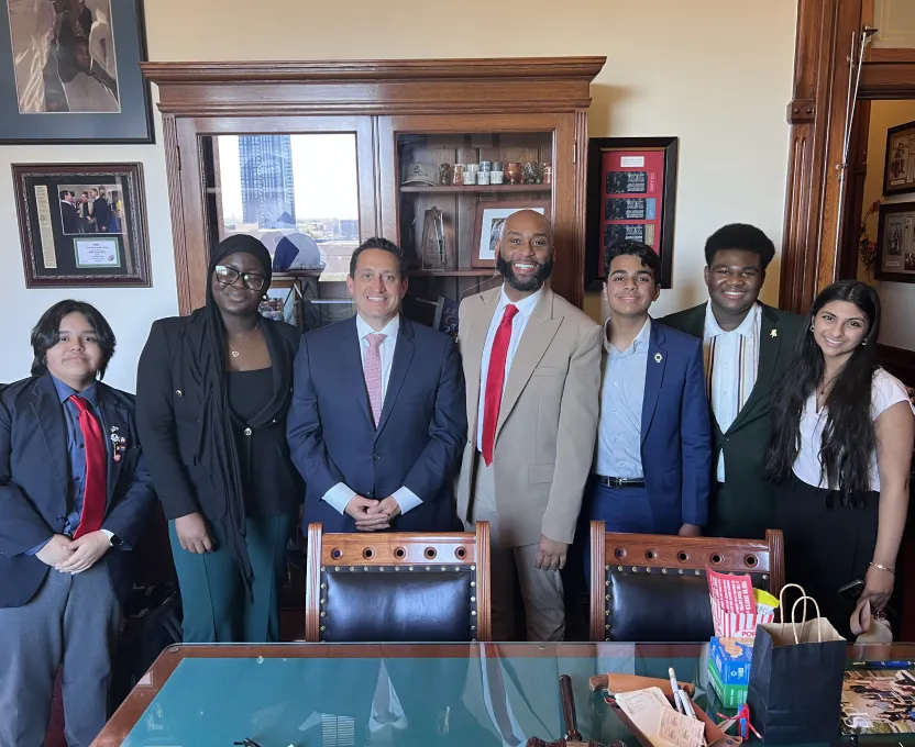 Group of six professionally dressed diverse people standing behind a desk in an office with wooden shelves and framed pictures on the wall.