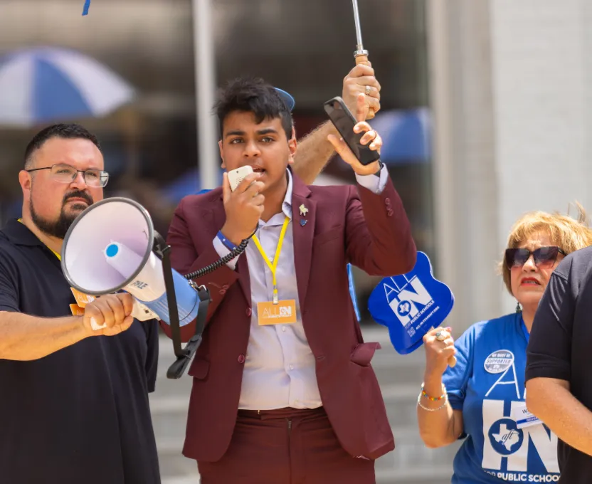 Young man in a burgundy suit speaking into a megaphone at a public demonstration, flanked by a man holding the megaphone and a woman holding a blue foam hand with 'ALL IN' text.