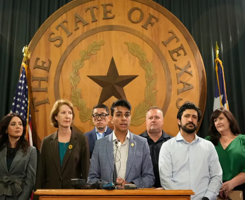 Group of six diverse people standing at a podium with microphones in front of a large wooden seal of The State of Texas and American flags on either side.