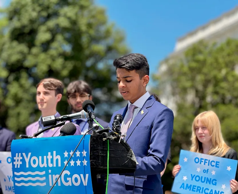 Teenage boy in a blue suit speaking at a podium with microphones and a #YouthinGov banner, with other youths standing behind holding signs.