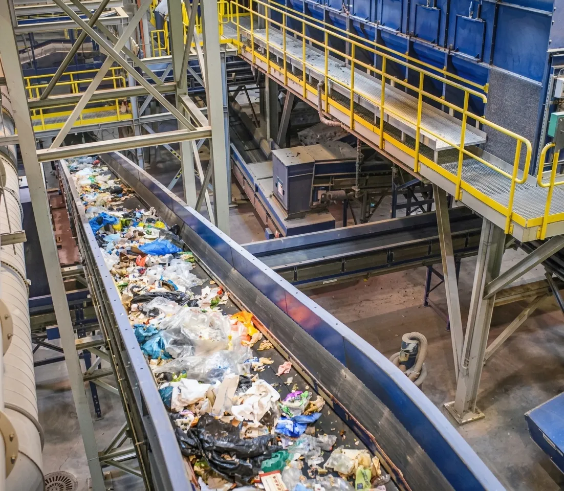 Conveyor belt in a recycling facility carrying mixed waste materials including plastic bags and packaging.