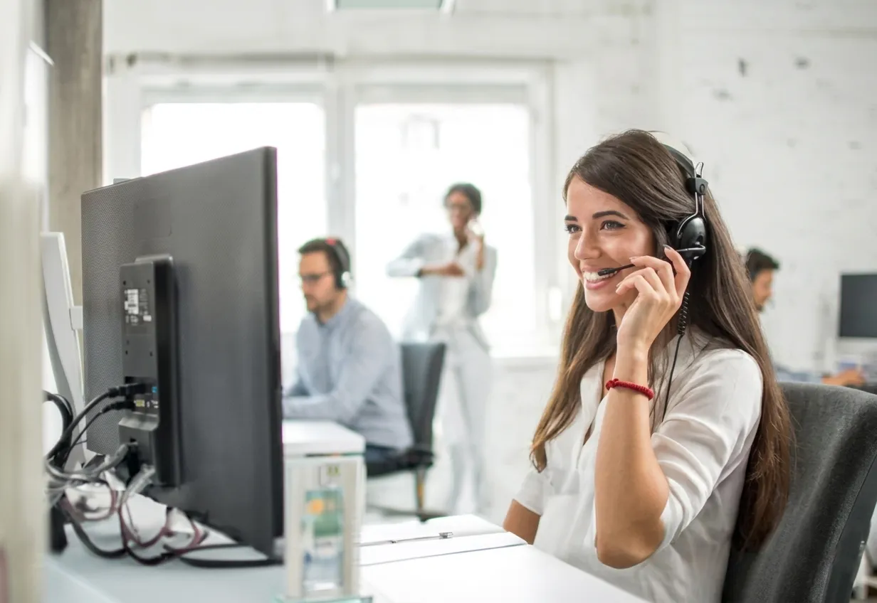 Smiling woman wearing headset working at computer in an office with colleagues in the background.