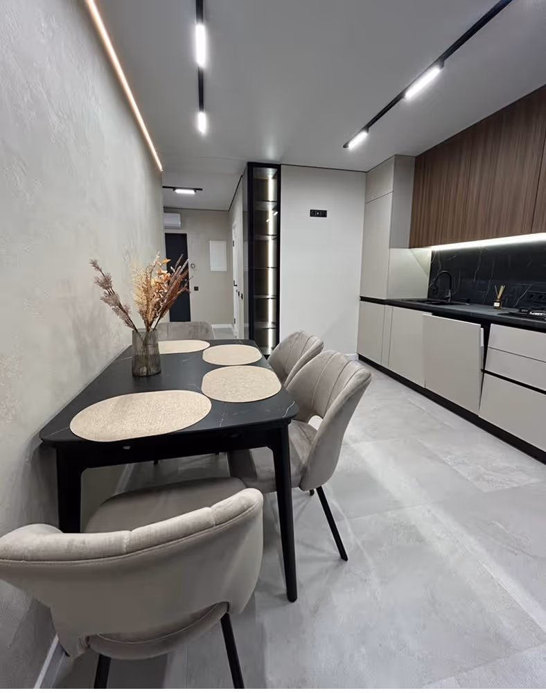 Сучасна кухня з чорним столом, чотирма світло-сірими стільцями, сухими квітами в прозорій вазі та білою і дерев'яною кухонною меблями.