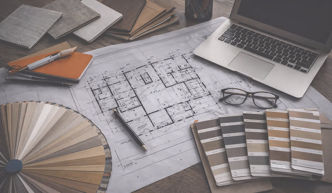 Архітектурний креслення будинку на столі з різними палітрами кольорів, ноутбуком, окулярами, блокнотами та ручками.