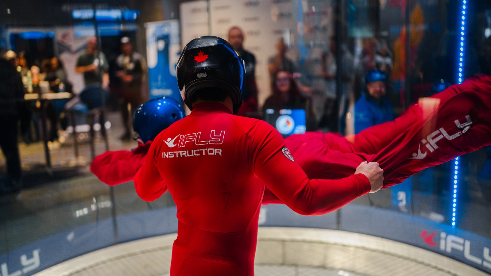 An iFLY instructor in red supporting a flyer inside the wind tunnel, shot through the glass