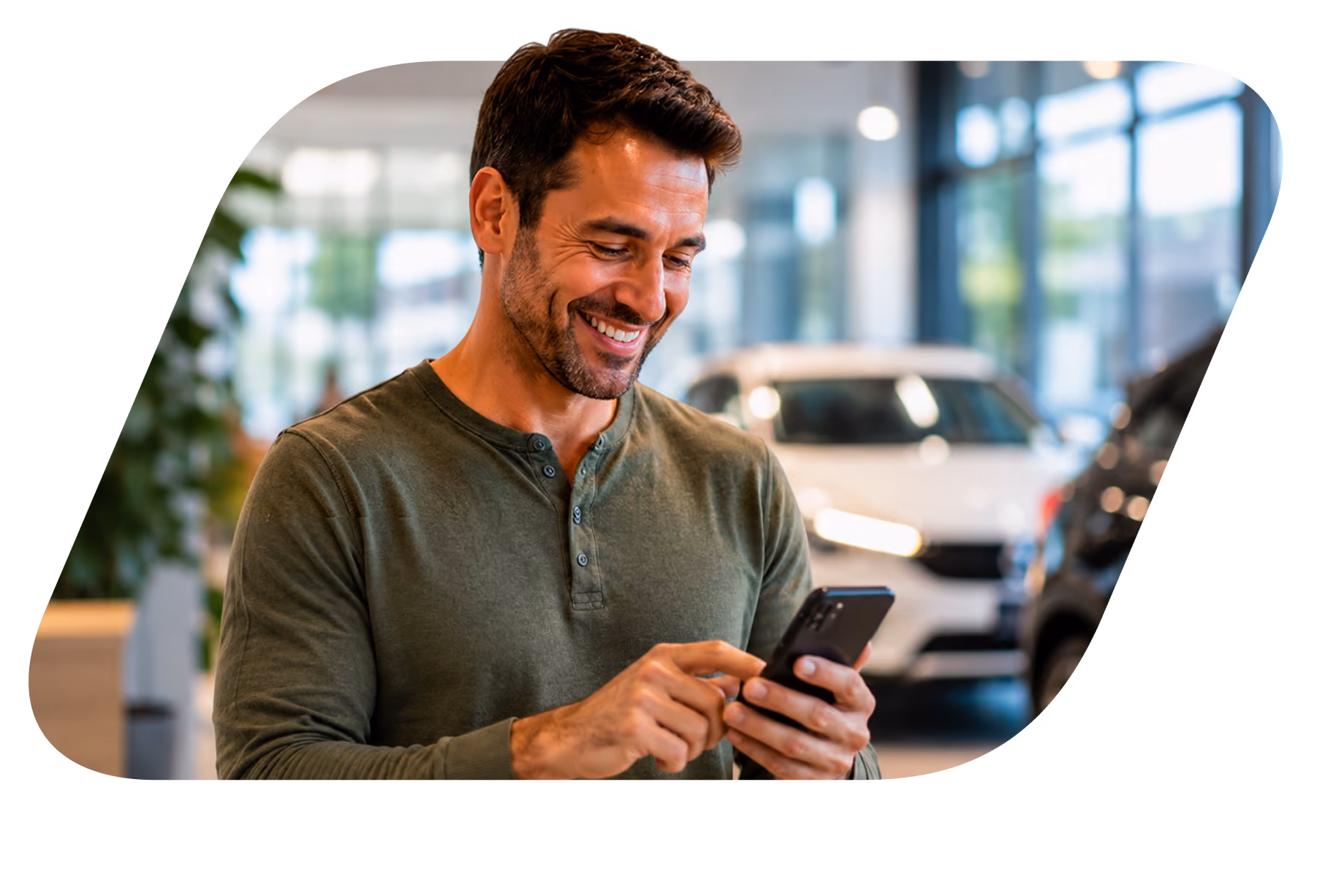 Homem sorridente usando smartphone em concessionária de carros com veículos ao fundo.