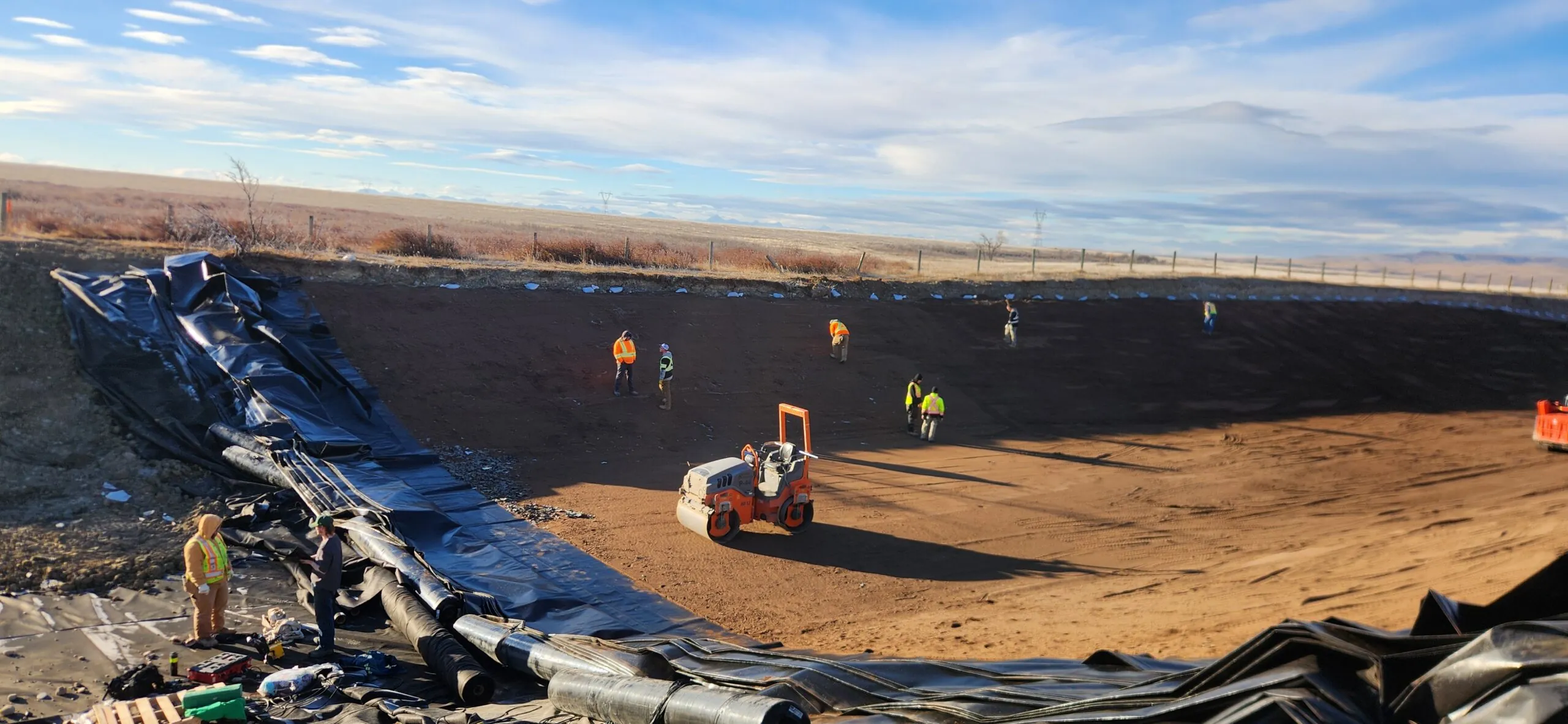 Workers in safety vests on a large dirt construction site with black liner and equipment.