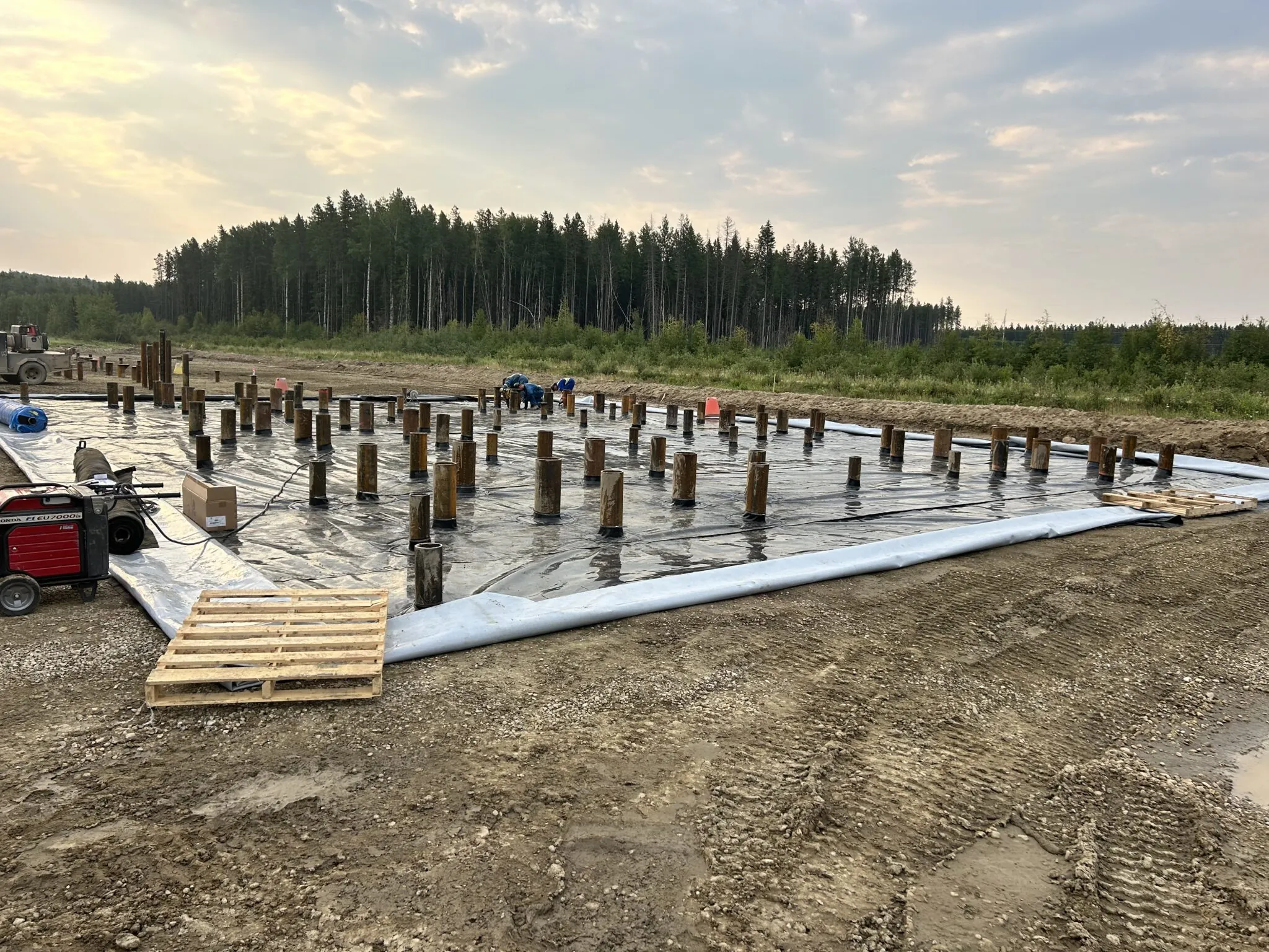 Construction site with metal pipes on plastic sheet laid on cleared dirt, surrounded by forest under cloudy sky.