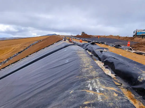 Workers installing large black plastic liners on a construction site under cloudy sky.