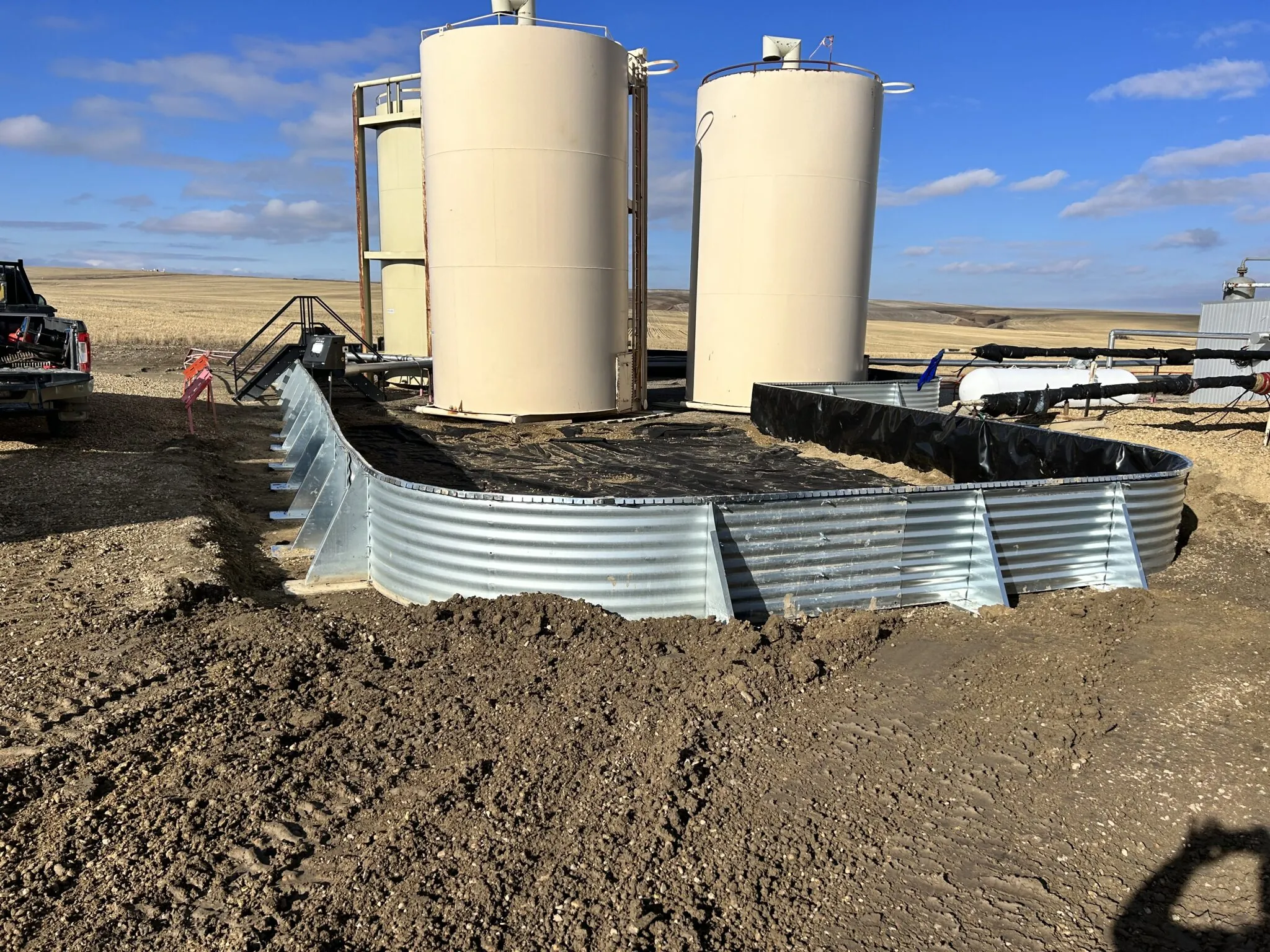 Two large beige storage tanks with a curved metal berm in front set on a dry dirt field under blue sky.