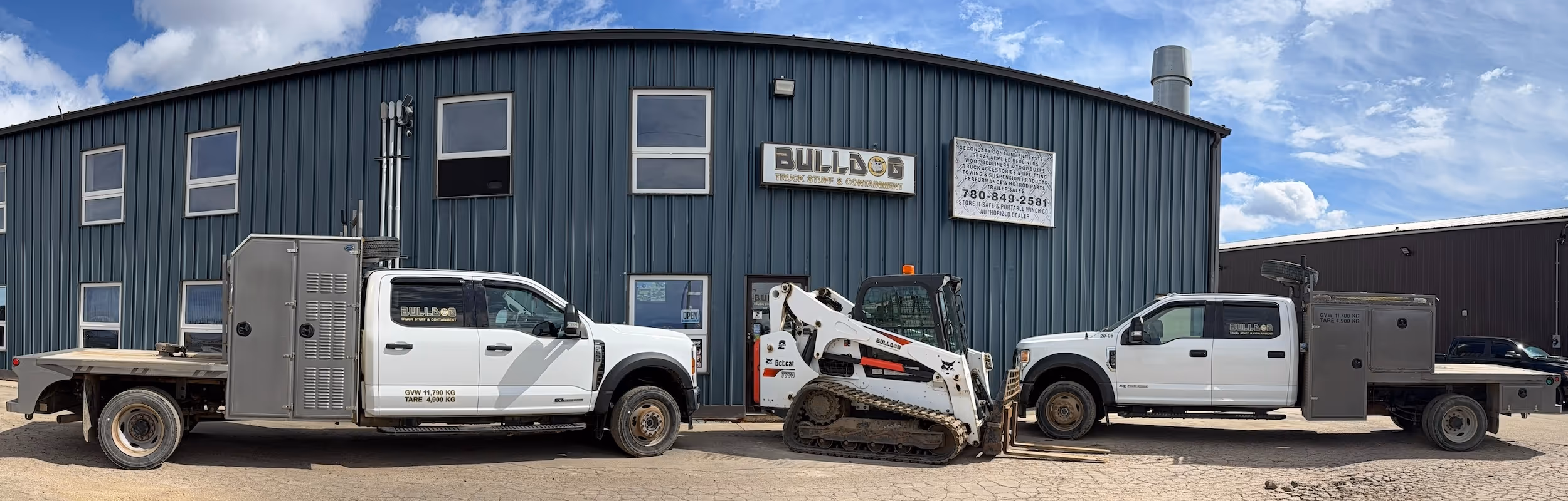 Two white flatbed trucks and a Bobcat skid-steer loader parked in front of a blue metal building.